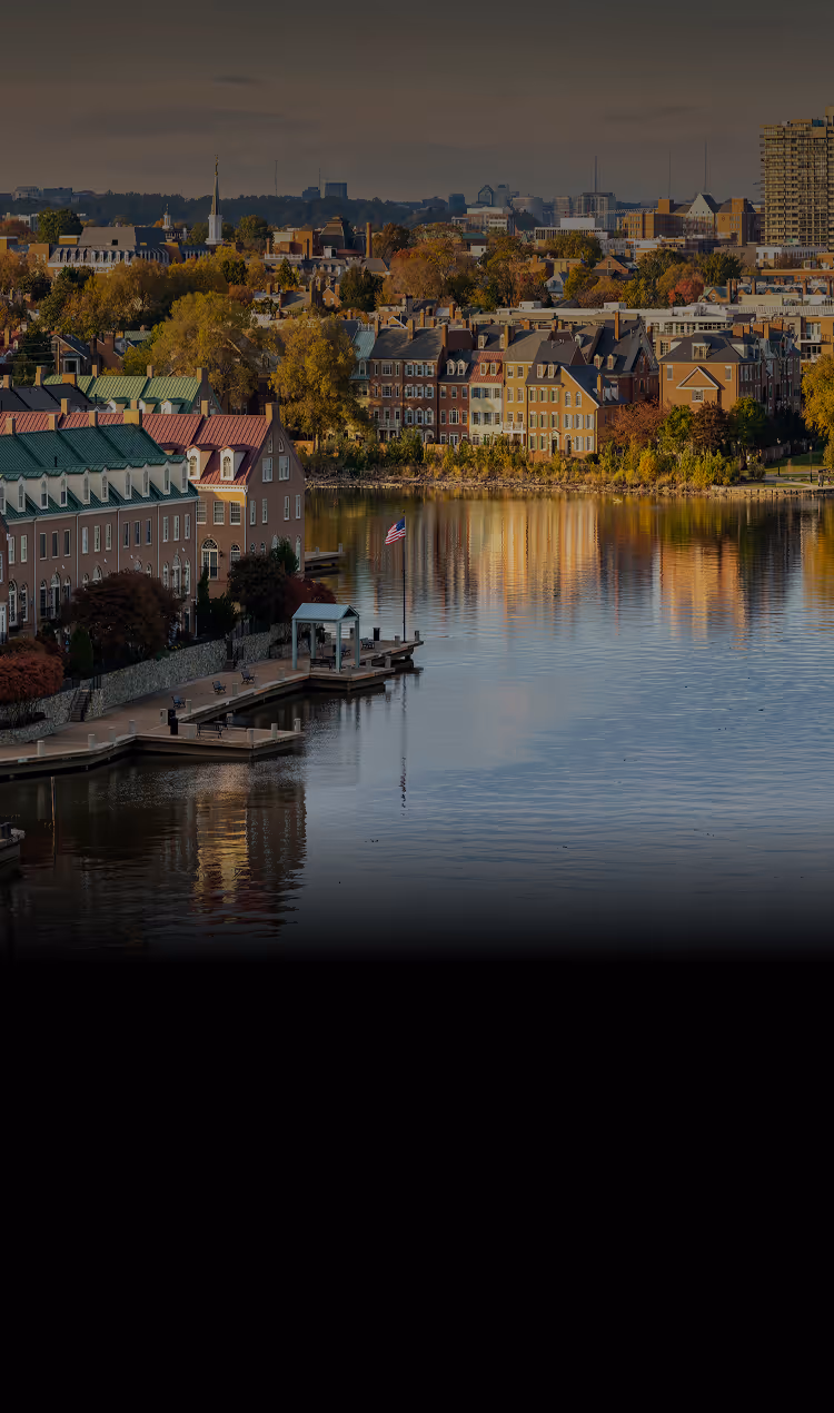 Waterfront view of historic Alexandria, Virginia with colonial-style buildings and an American flag on a dock reflecting in the calm water.