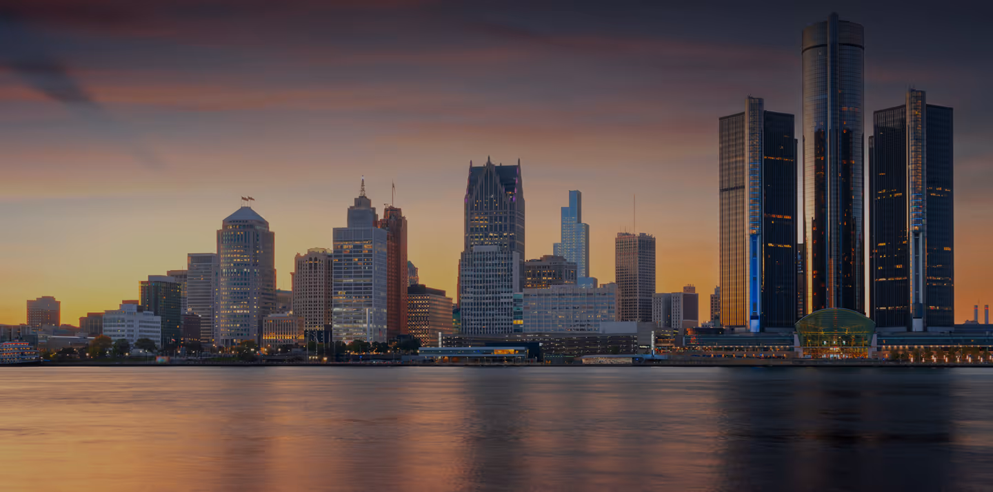 Detroit skyline at sunset with tall buildings reflecting on the Detroit River.