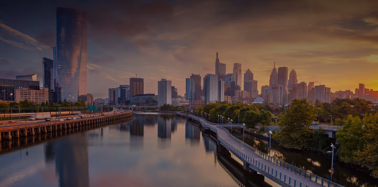 Philadelphia city skyline at sunset with river and pedestrian bridge in the foreground.
