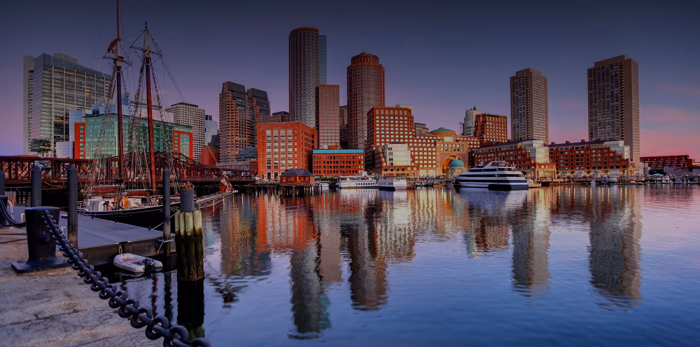 Boston skyline at dusk reflected in calm harbor water with boats docked in foreground.