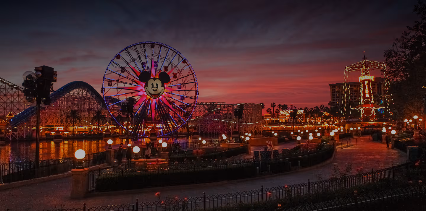Illuminated Mickey Mouse Ferris wheel and roller coaster at a Disney theme park during sunset with a vibrant red and purple sky.