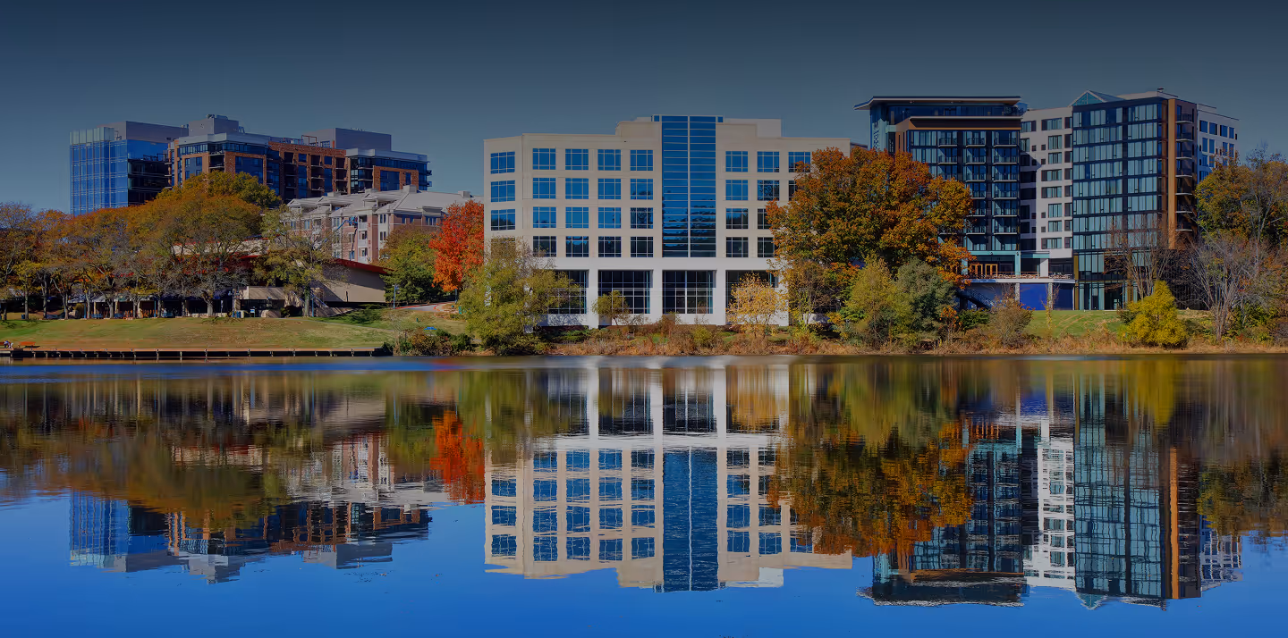 Modern office buildings and autumn trees reflecting on the calm water of a lake under clear blue sky.