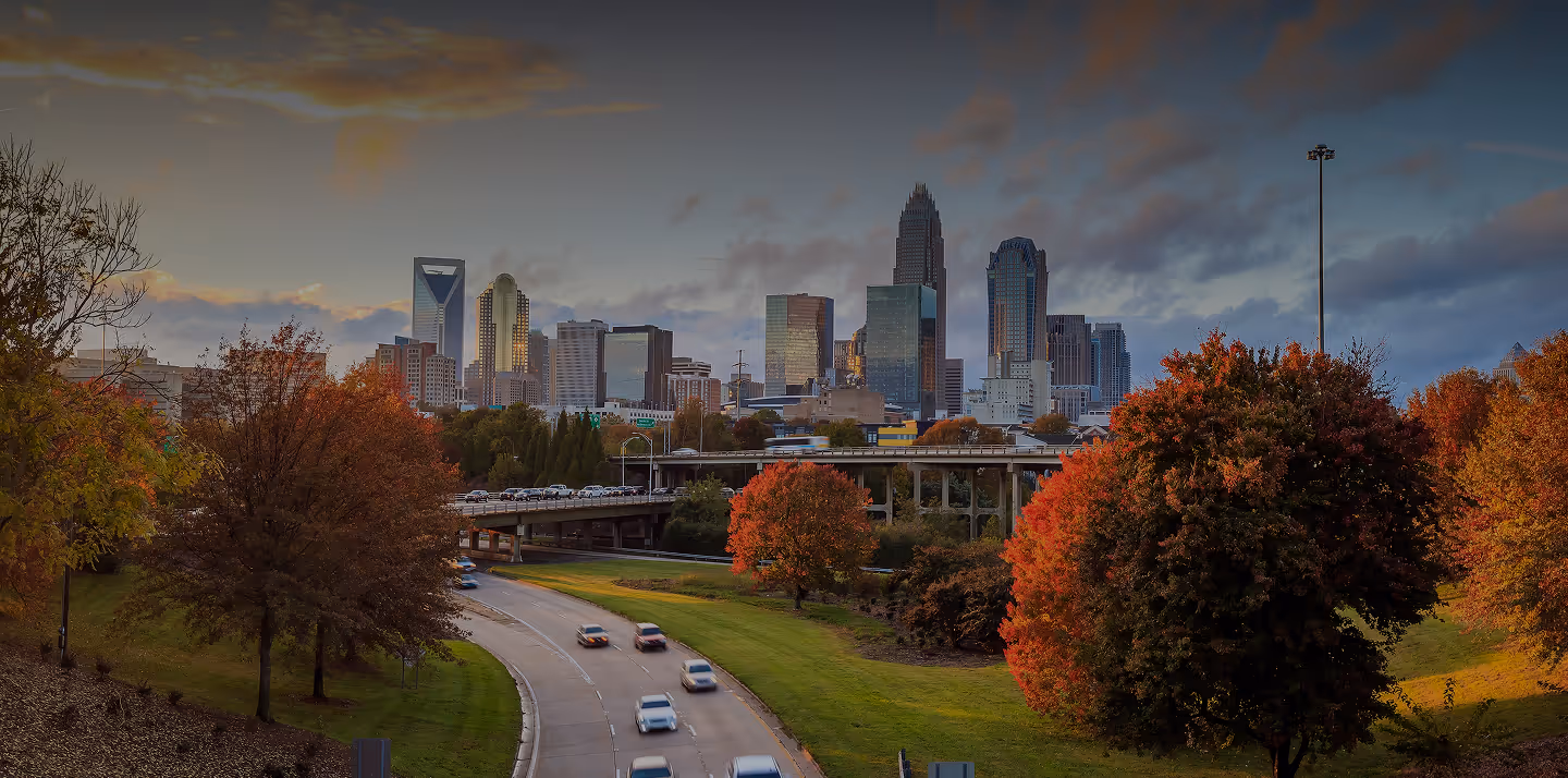 Wide view of Charlotte skyline at sunset with autumn trees and cars on a curved highway.