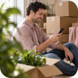 A man booking a move on his phone surrounded by moving boxes