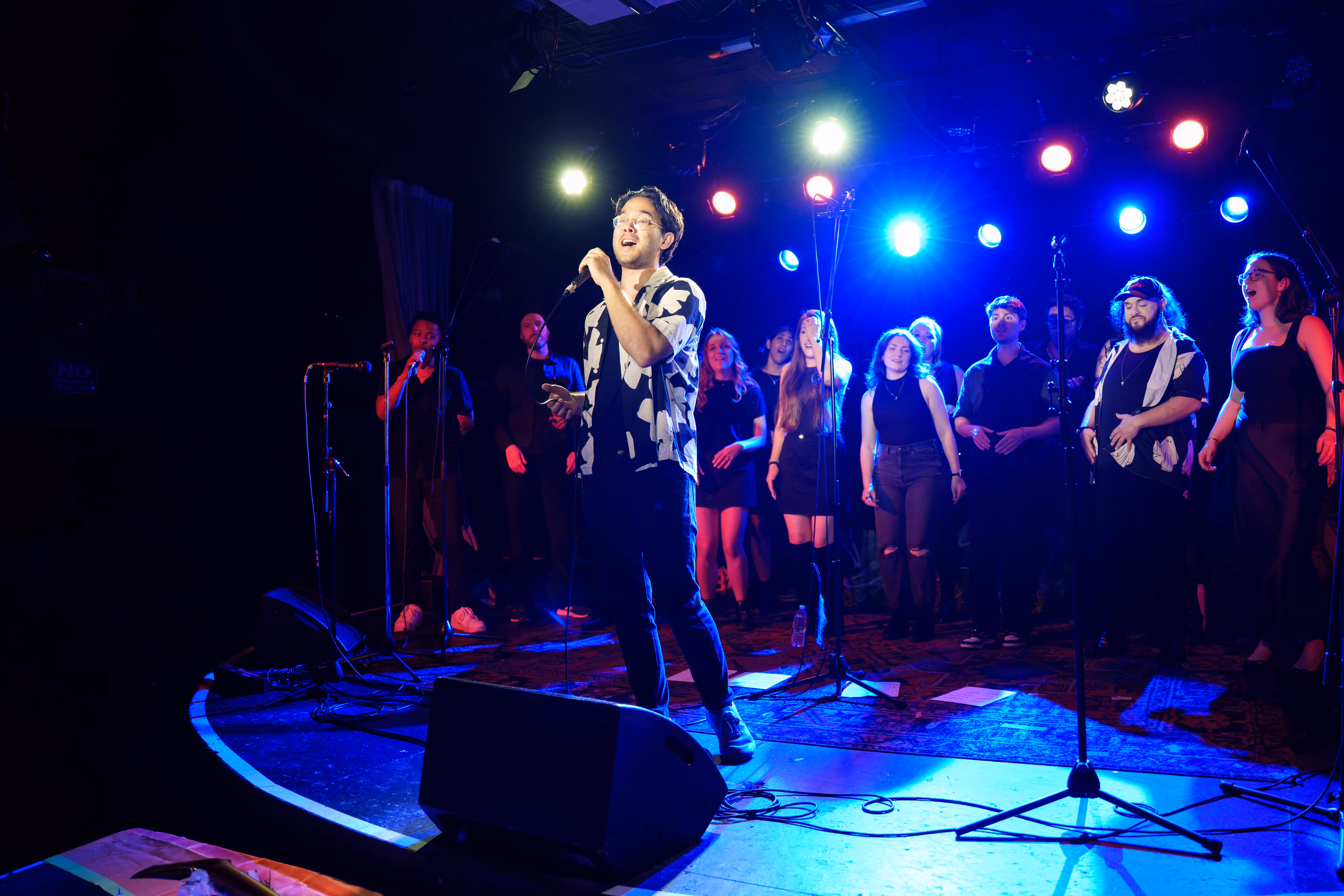 Man wearing glasses and a patterned shirt holding a microphone on stage under purple and white lights with three people blurred in the background.