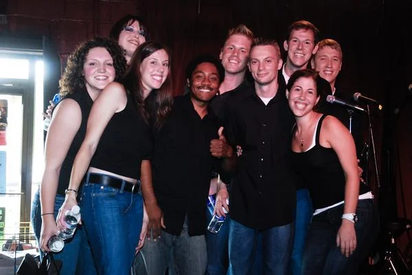 Group of eight young adults smiling and posing together indoors near microphones, some holding water bottles.