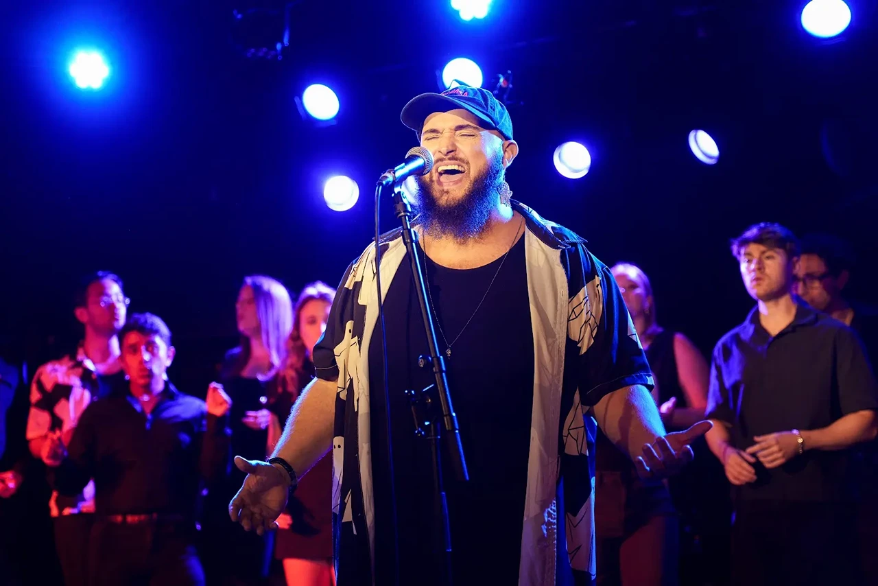 Bearded male singer passionately performing on stage with backup singers and blue stage lights overhead.