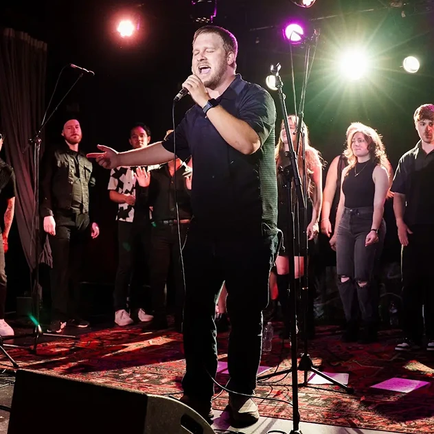 Male singer passionately performing with a microphone on stage, accompanied by a group of vocalists in the background.