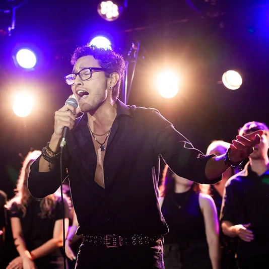 Man with glasses passionately singing into a microphone on stage with backup singers and bright stage lights behind him.