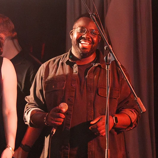 Smiling man wearing glasses and a dark shirt holding a microphone on stage with a microphone stand.