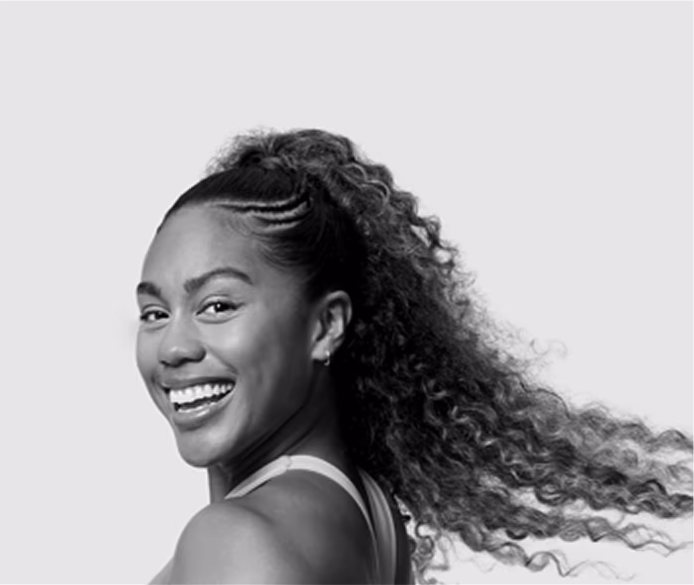 Smiling woman with curly hair tied back and wearing a sleeveless top against a plain background.