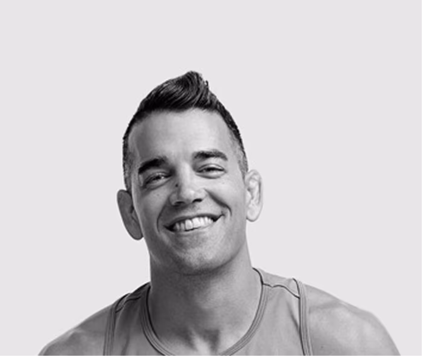 Smiling man with short hair styled upwards wearing a sleeveless top against a plain background.