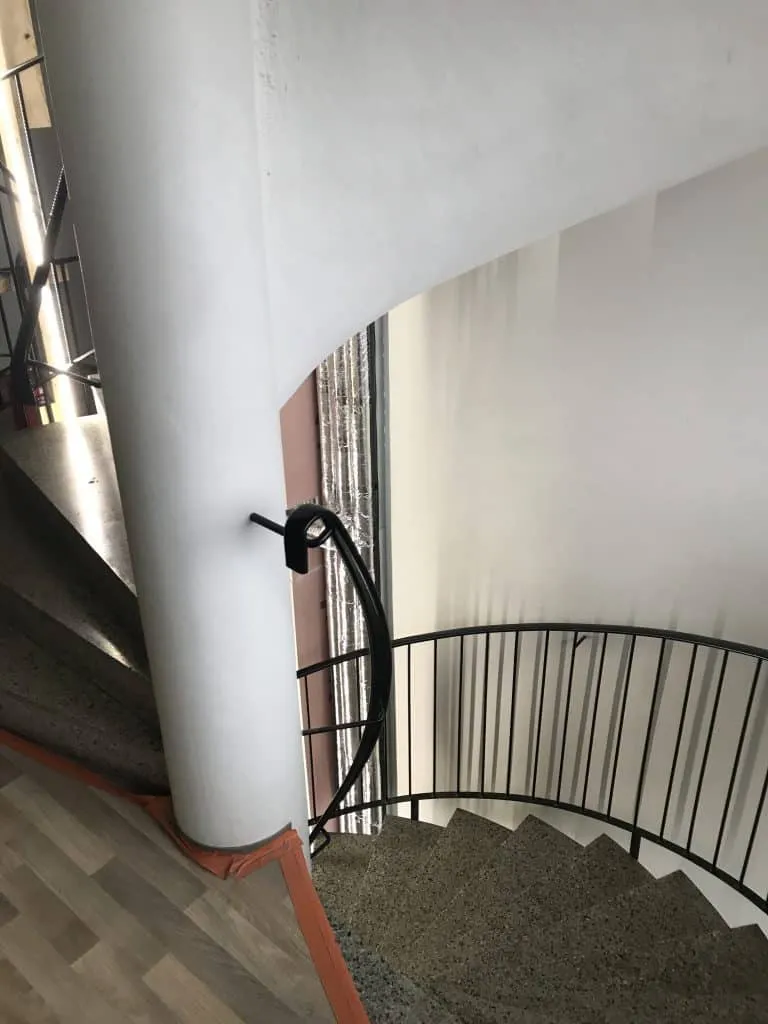 Ylhäältä kuvattu kierreportaikko, jossa on valkoinen pylväs, musta kaide ja harmaat kiviset portaat. Lattialla on puujäljitelmäpintaa ja oranssi reunus.