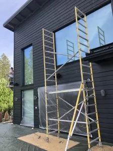 Tumman ulkoverhouksen omaava talo, jonka edessä on kelta-hopeinen rakennusteline ja suojamuovi peittämässä ovea. Taustalla näkyy vihreää kasvillisuutta ja sininen taivas.