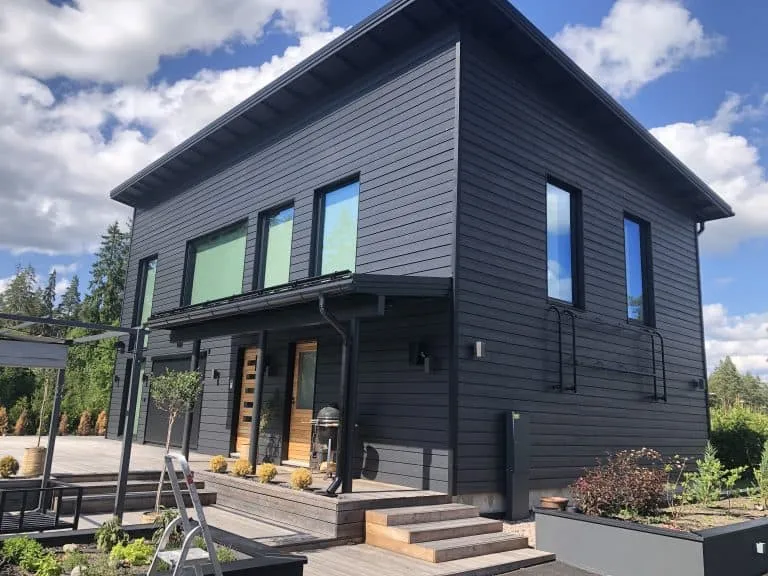Moderni, tummanharmaa kaksikerroksinen talo puuverhoilulla, jossa on suuret ikkunat ja puinen terassi, valoisassa säässä.