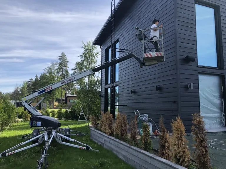 Ulkona on nosturi, jolla maalataan tummaa puutaloa, jossa on suuret ikkunat ja mies korissa maalaamassa.