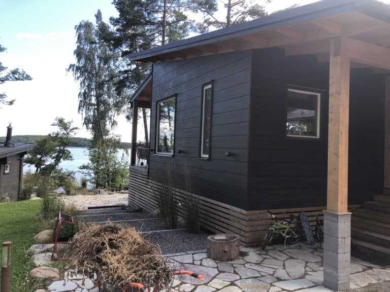 Moderni, tumma puutalo järven rannalla, jossa on suuret ikkunat ja kivipolku. Aurinkoinen sää valaisee maisemaa.