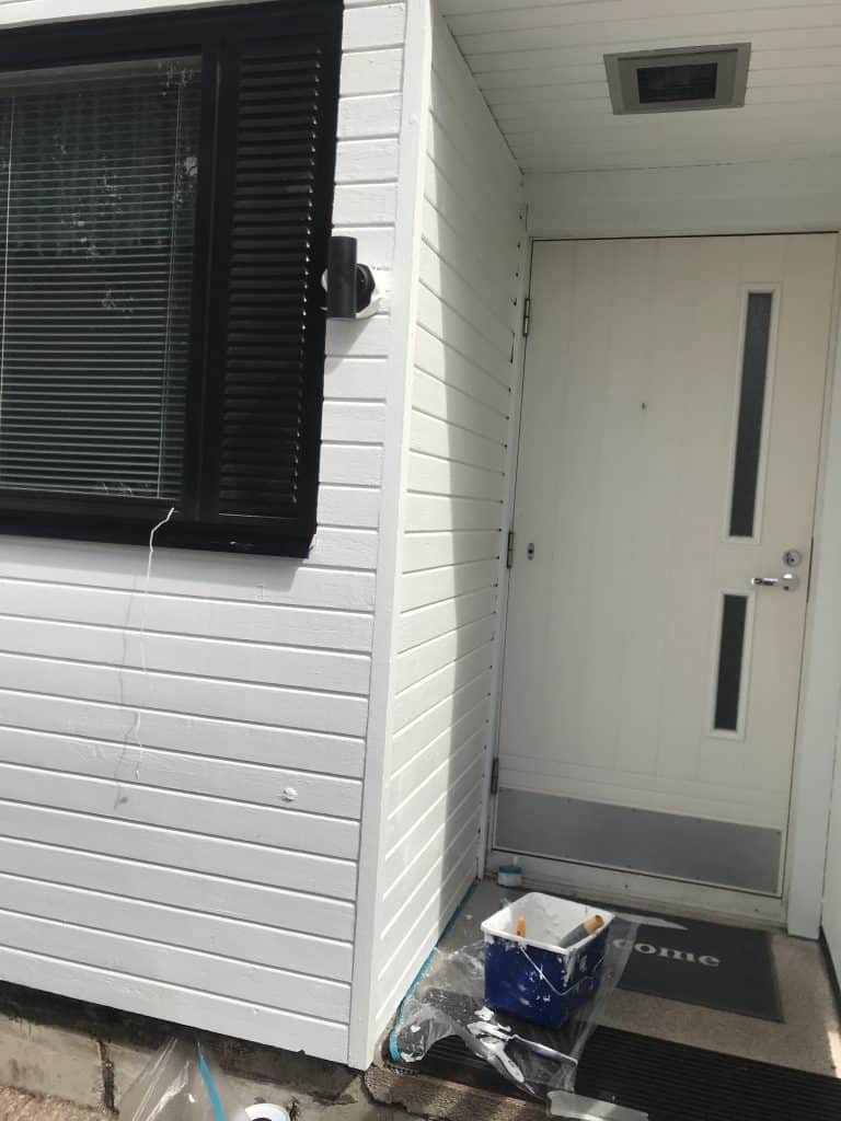 Ulkokuva valkoisesta puutalosta, jossa on musta ikkuna ja ovi, sekä maalaustarvikkeita.