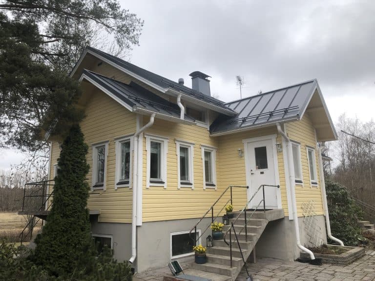 Kaksikerroksinen keltainen puutalo harmaalla katolla, portaat johtavat etuovelle, pilvinen sää.