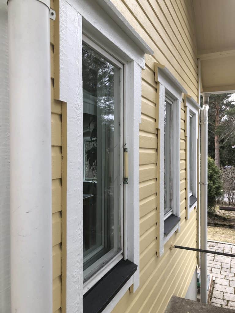 Vaaleankeltaisen puutalon ulkoseinä, jossa on valkoiset ikkunankarmit ja valkoinen sadevesiputki. Ikkunat heijastavat ulkoilmaa.