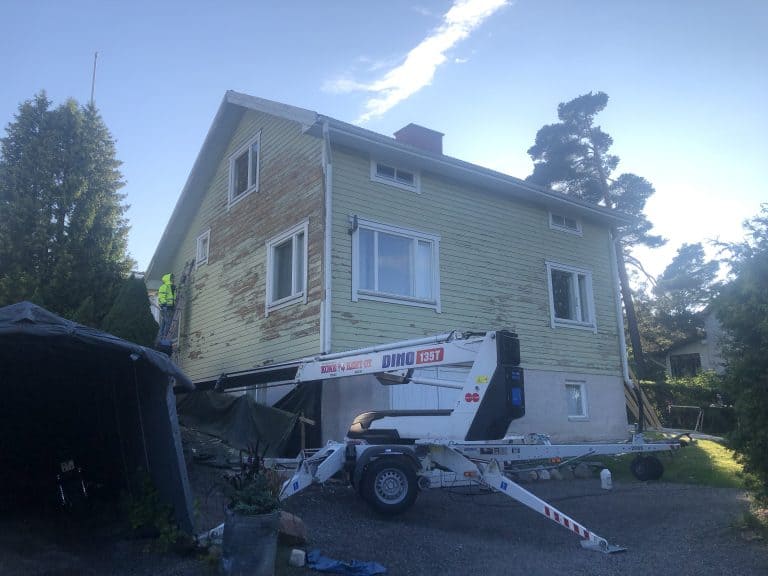 Vaaleanvihreä puutalo, jossa on irtoavaa maalia, nostolava ja työntekijä, aurinkoinen sää.