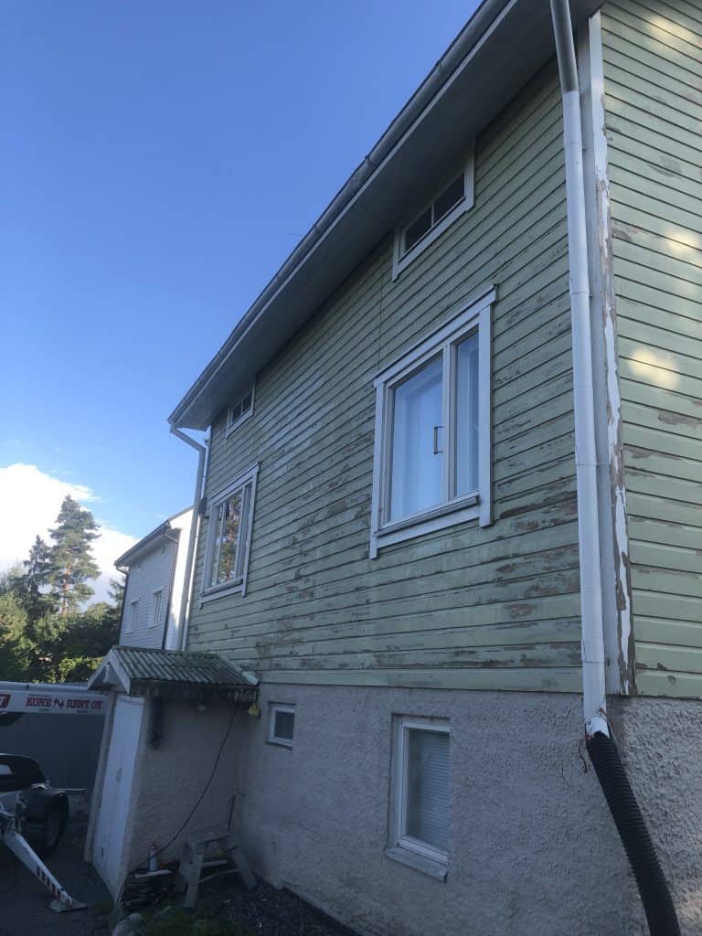 Kaksikerroksinen puutalo, jossa on vihreä ulkovuoraus ja valkoiset ikkunakehykset. Maali on hilseilemässä. Alakerrassa on harmaa kivijalka. Taustalla näkyy sininen taivas ja puita.