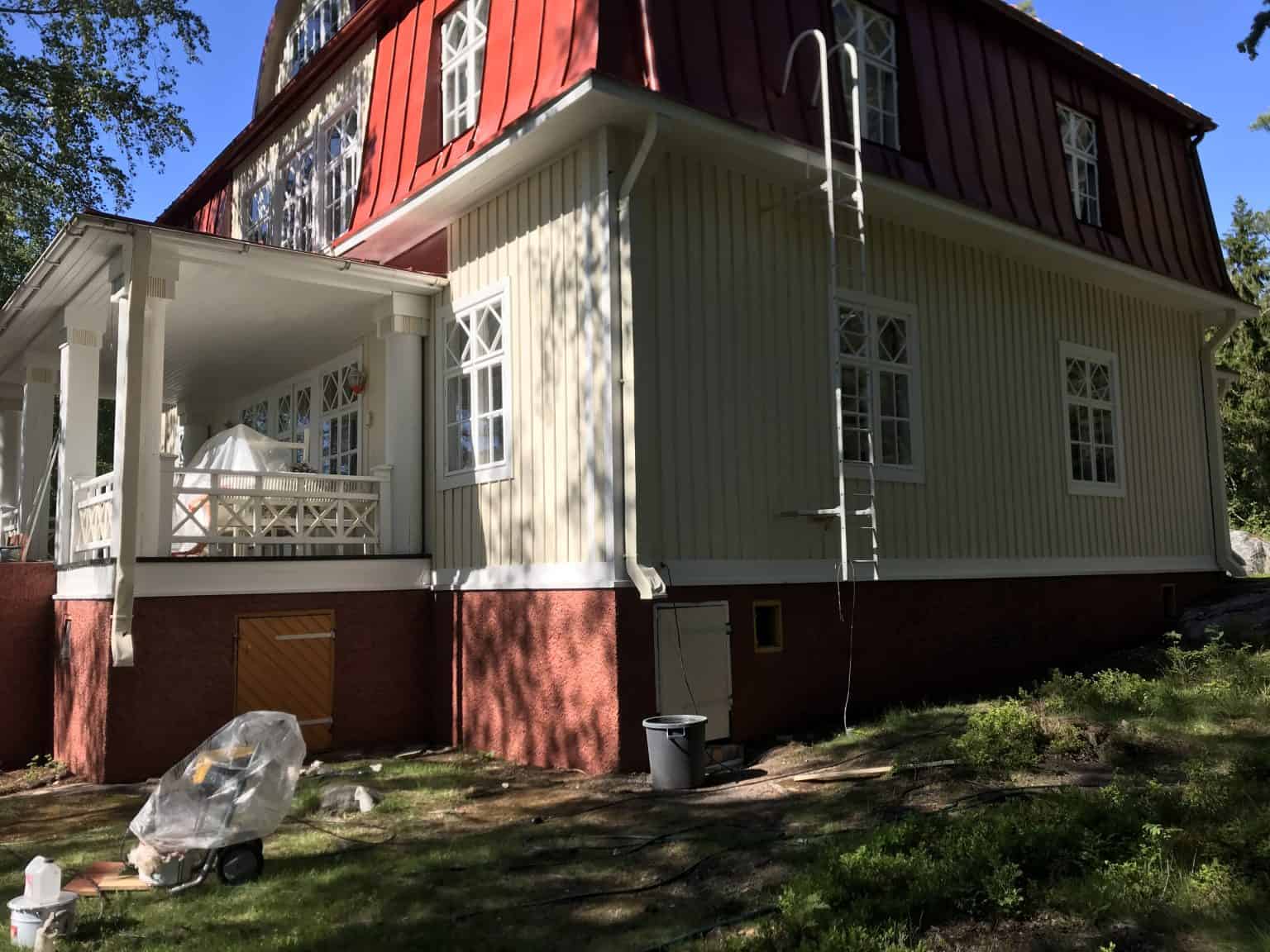 Vaaleankeltainen puutalo punaisella katolla, valkoiset ikkunat ja pylväät, aurinkoinen sää.