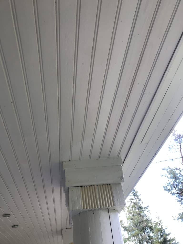 Valkoinen puinen pylväs tukee valkoista paneelikattoa, ulkona näkyy puita ja taivasta.