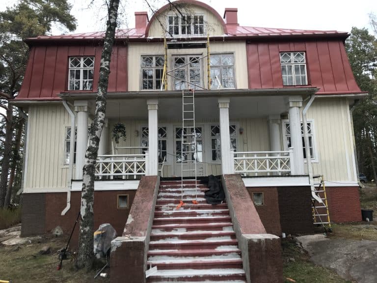 Kaksikerroksinen, vaaleankeltainen talo punaisella katolla, jossa on valkoiset pylväät ja portaat. Etupihalla on puu ja kaksi tikapuut.