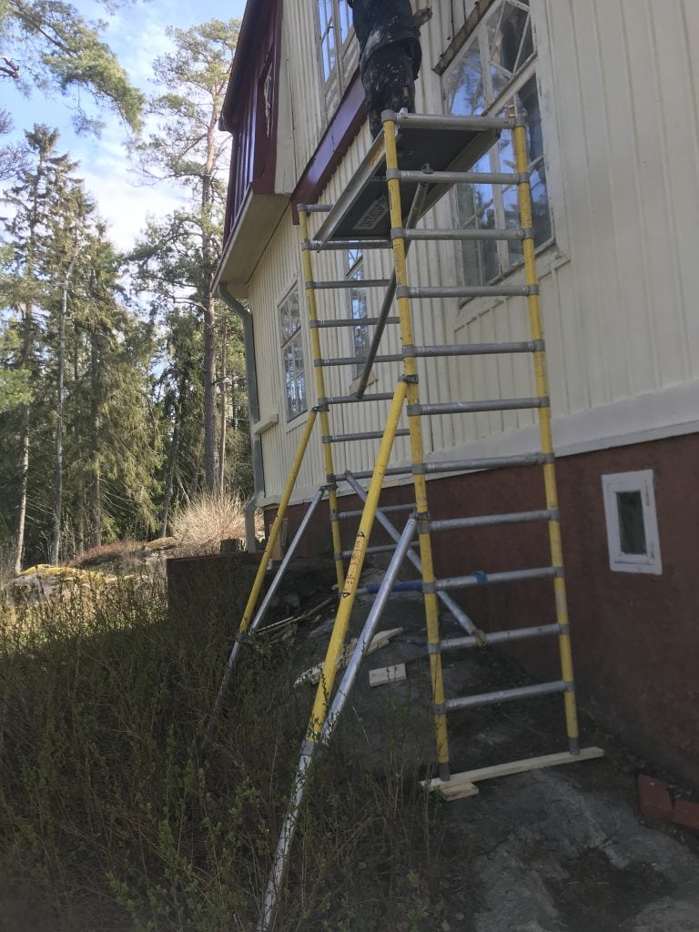 Ulkona on rakennustelineet talon seinustalla, jossa henkilö maalaa. Telineet ovat keltaiset ja hopeanväriset, ja talo on maalattu vaaleankeltaiseksi punaisella pohjalla.