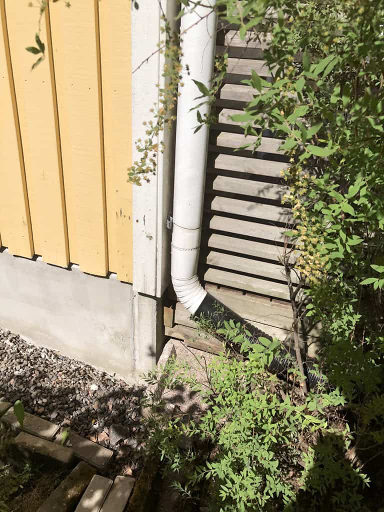 Ulkokuva, jossa näkyy keltainen puutalo, valkoinen syöksytorvi, harmaa kivijalka, kivet ja vihreitä kasveja aurinkoisessa säässä.