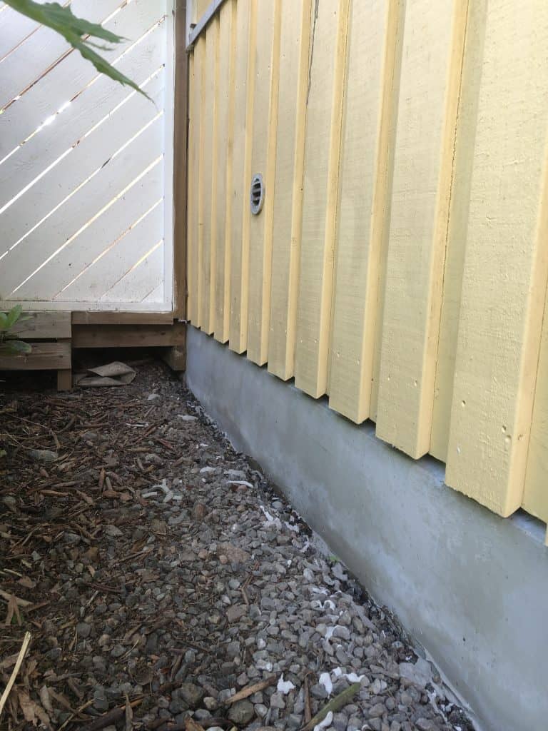 Kuva näyttää ulkokuvaa keltaisesta puutalosta, jossa on harmaa kivijalka ja valkoinen aita vasemmalla. Maassa on kiviä ja risuja. Valo on kirkas ja luonnollinen.