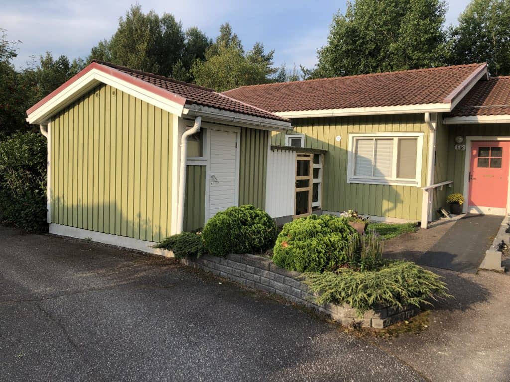 Vaaleanvihreä puutalo, jossa punainen katto, valkoiset ikkunat ja ovi, sekä vihreitä pensaita ja asfalttipiha.