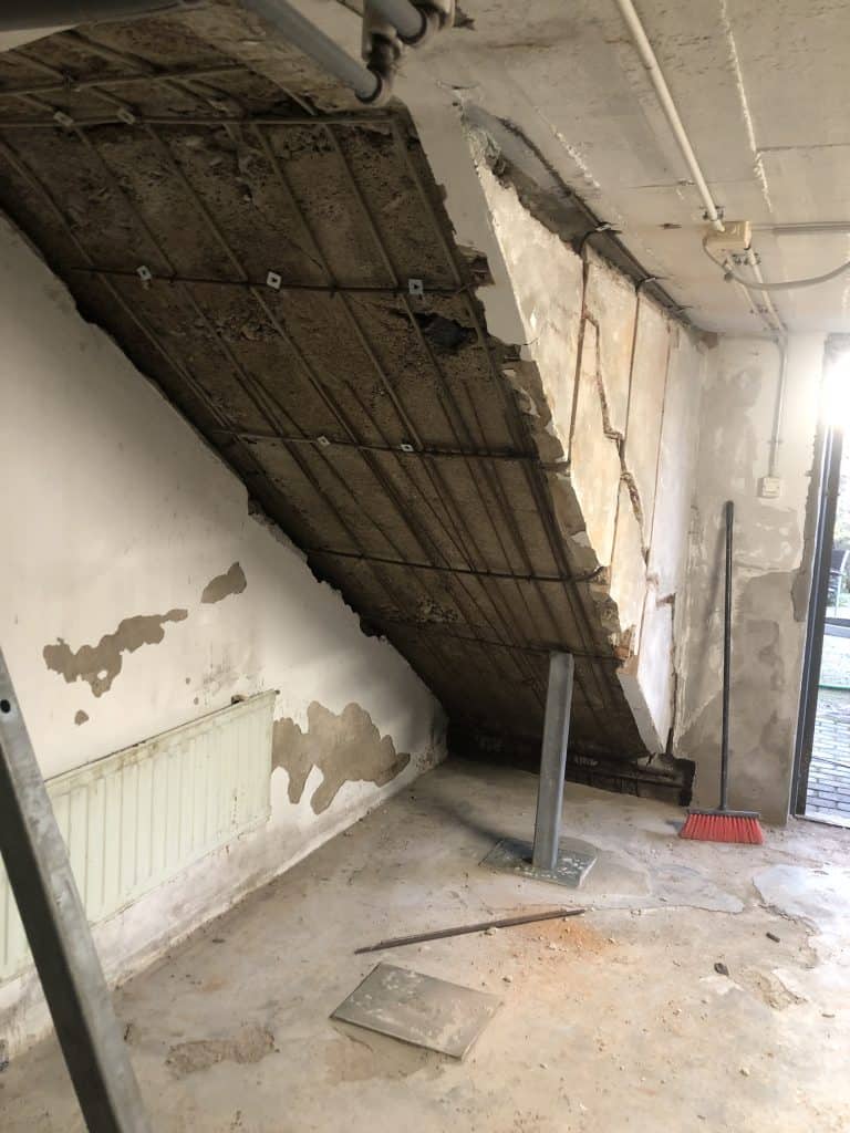 Huoneessa on vaurioitunut katto, jossa näkyy raudoitus. Seinät ovat rapistuneet ja lattialla on rakennusjätettä. Huoneessa on myös patteri, tukipylväs ja harja.