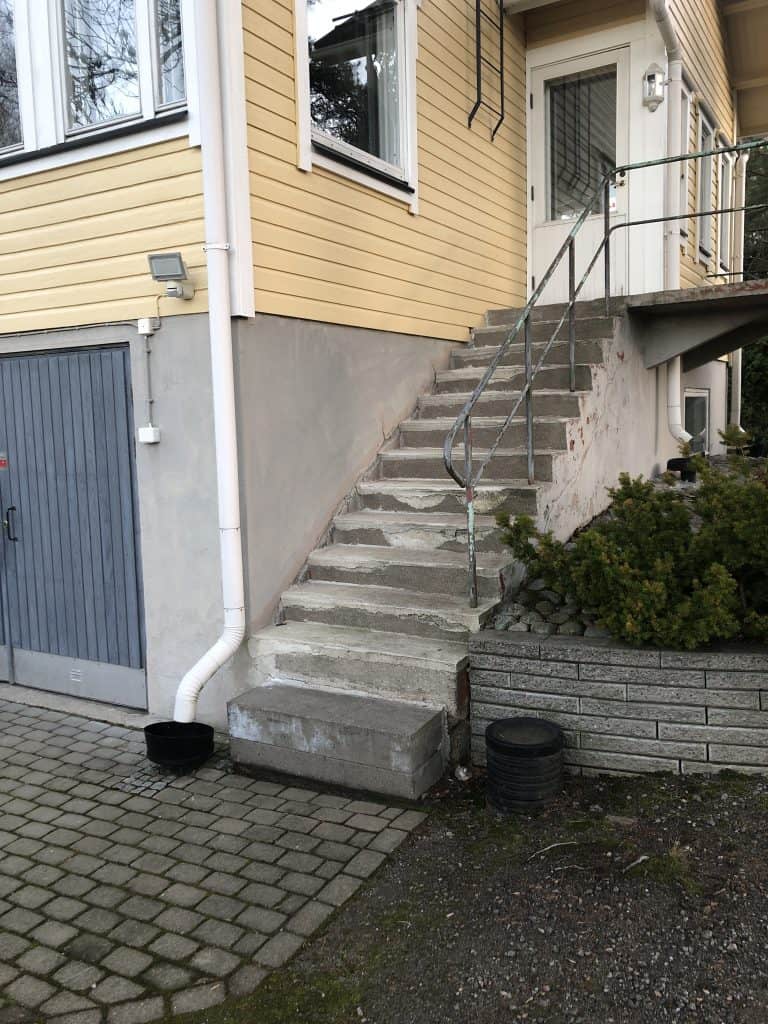 Ulkokuva keltaisesta talosta, jossa on harmaat portaat, harmaa autotallin ovi ja vihreitä pensaita.