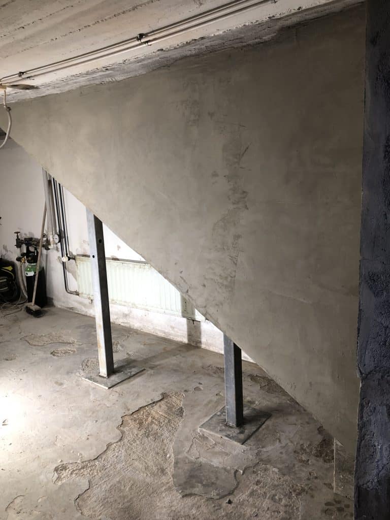 Kuva näyttää betonisen portaikon, jota tukevat metallipylväät, karkean betonilattian ja valkoisen patterin taustalla, valaistus on himmeä.