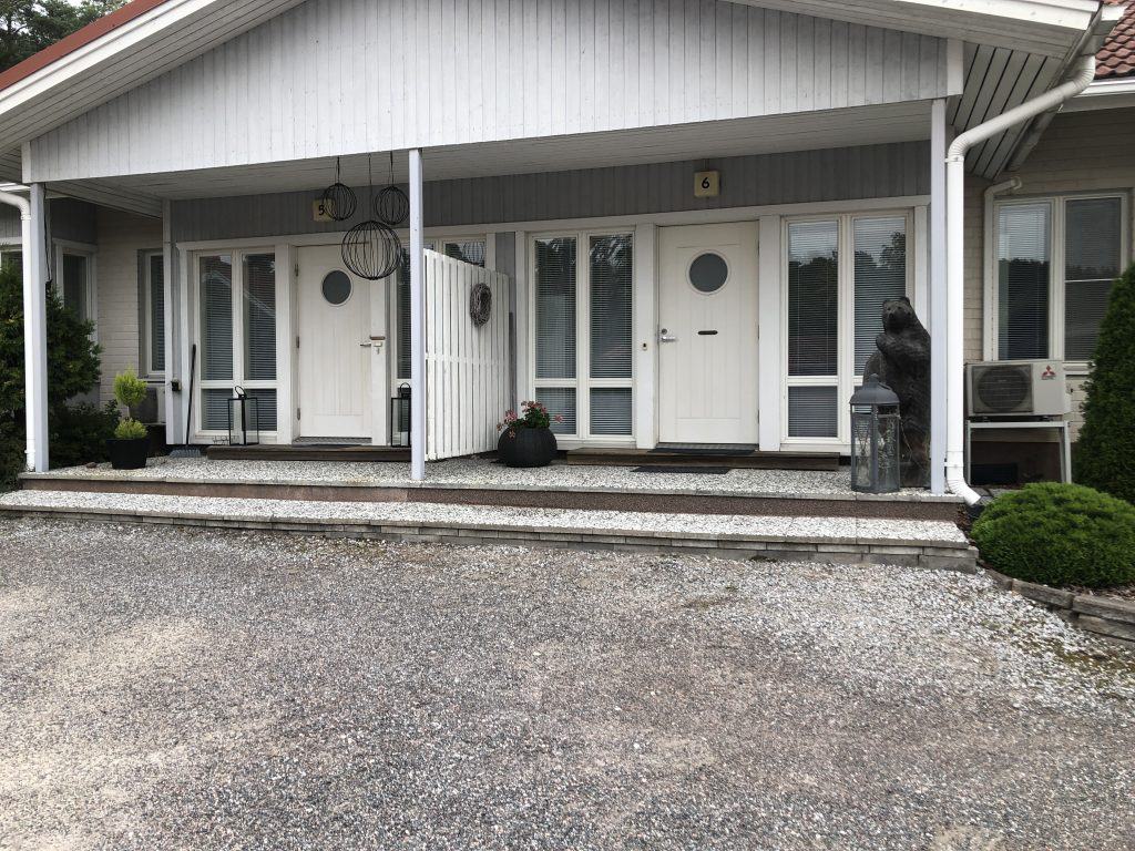 Rivitalon julkisivu, jossa on kaksi valkoista ovea pyöreillä ikkunoilla, numeroilla 5 ja 6 merkittyinä, sekä koristeellisia lyhtyjä ja kasveja.