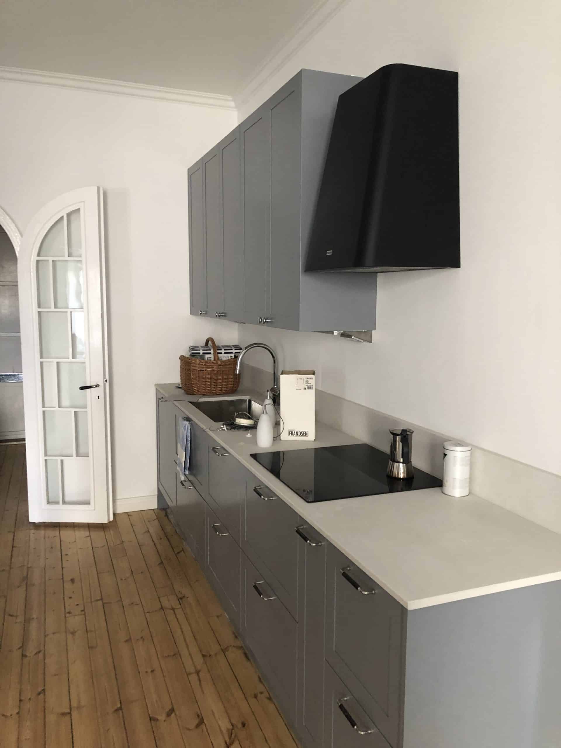 Vaaleanharmaassa keittiössä on puulattia, valkoiset seinät, harmaat kaapit, musta liesituuletin ja valkoinen työtaso. Korissa on astioita ja tiskialtaan vieressä on Frandsen-laatikko.