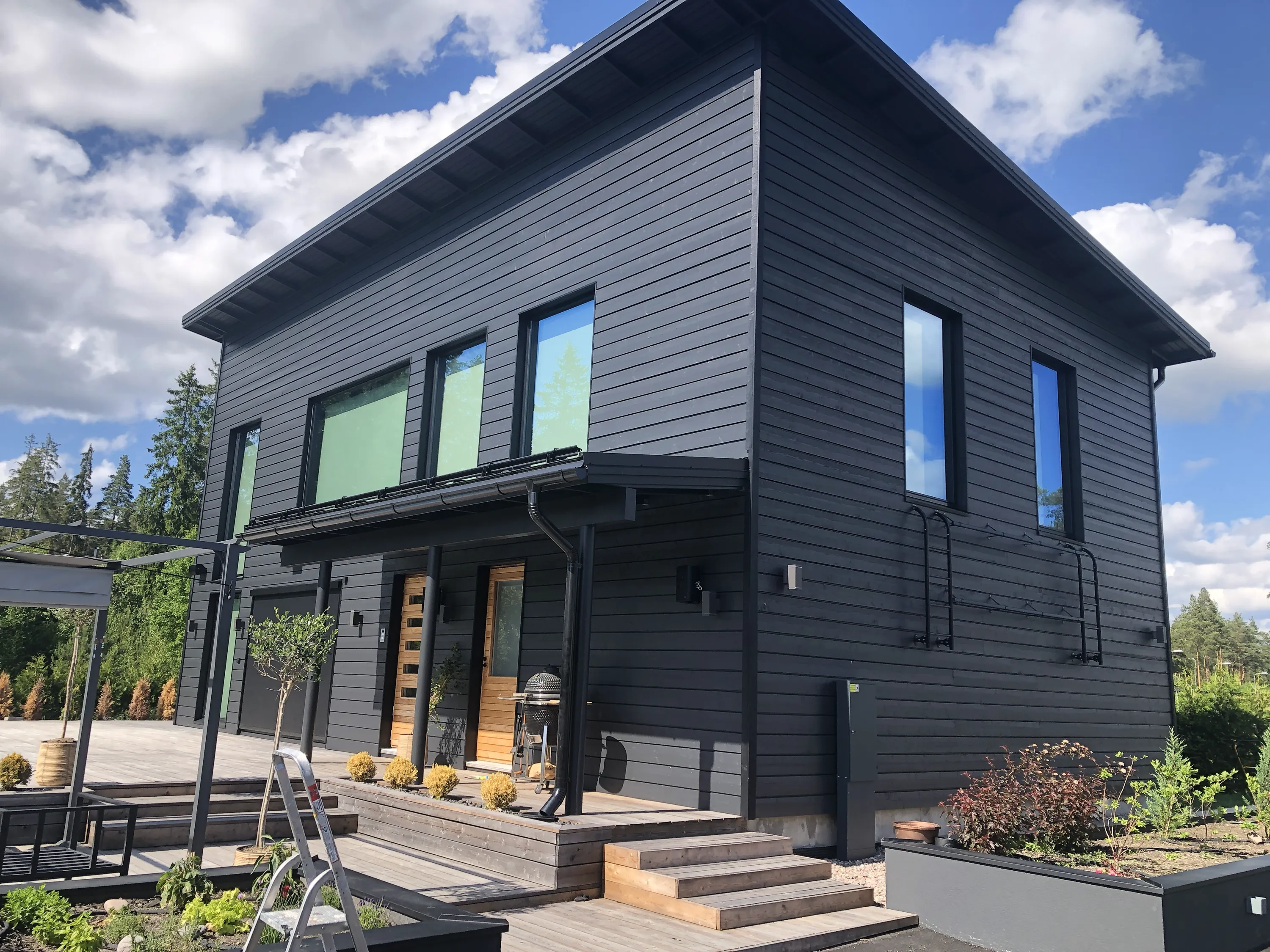 Moderni, tummanharmaa puutalo, jossa on suuret ikkunat ja puinen terassi, aurinkoisena päivänä.