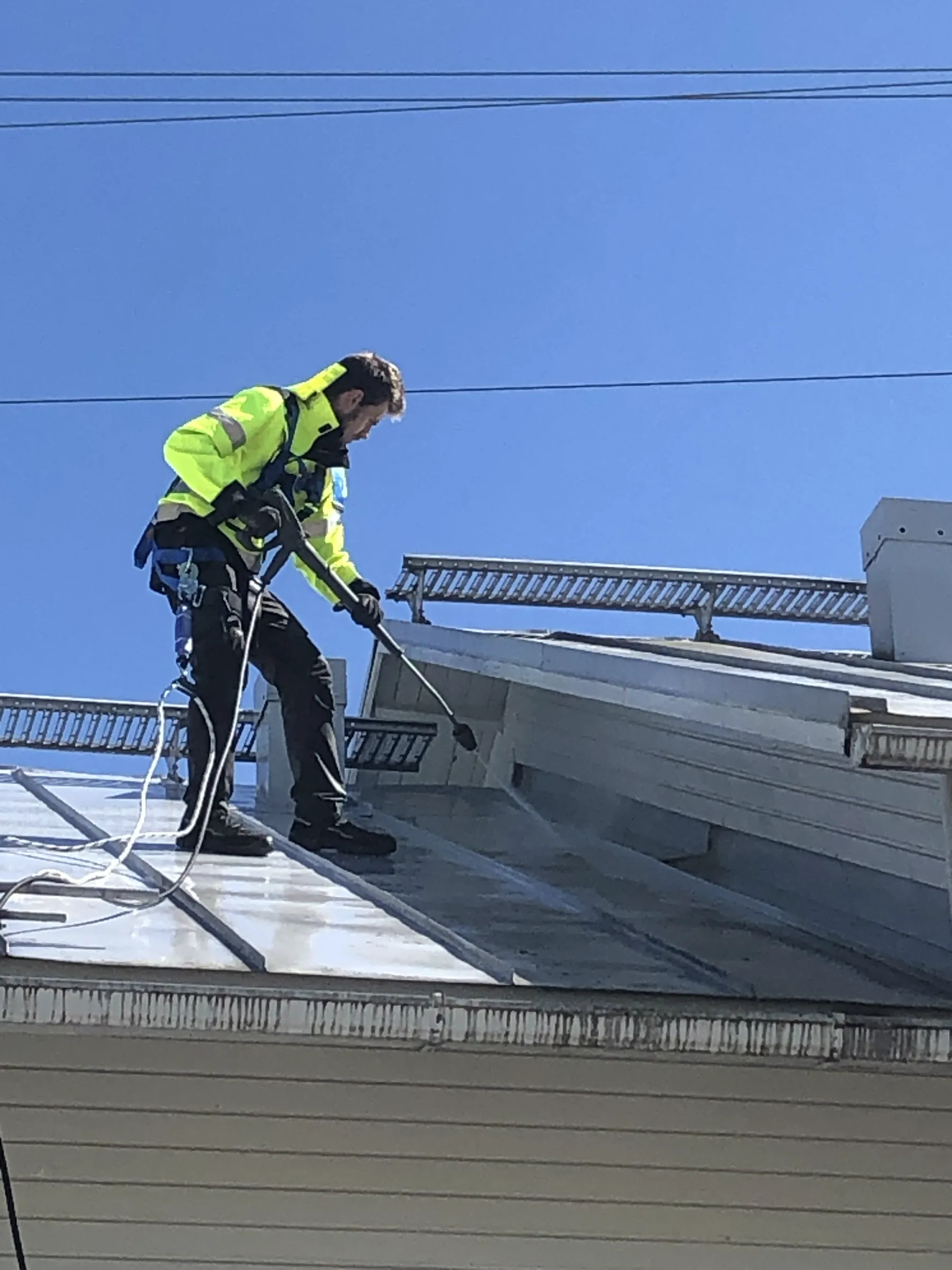 Mies painepesee kattoa, yllään suojavarusteet ja kirkkaan keltainen takki, aurinkoisessa säässä.