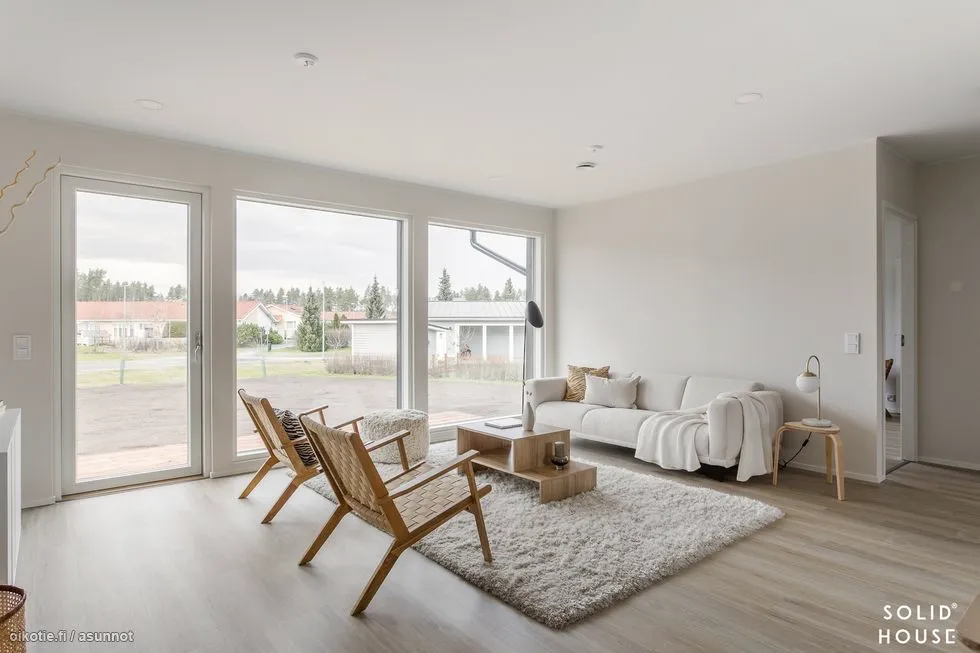 Valoisassa olohuoneessa on suuret ikkunat, vaalea sohva, puinen sohvapöytä, kaksi rottinkituolia ja pehmeä matto. Huoneessa on myös jalkalamppu ja pieni sivupöytä.