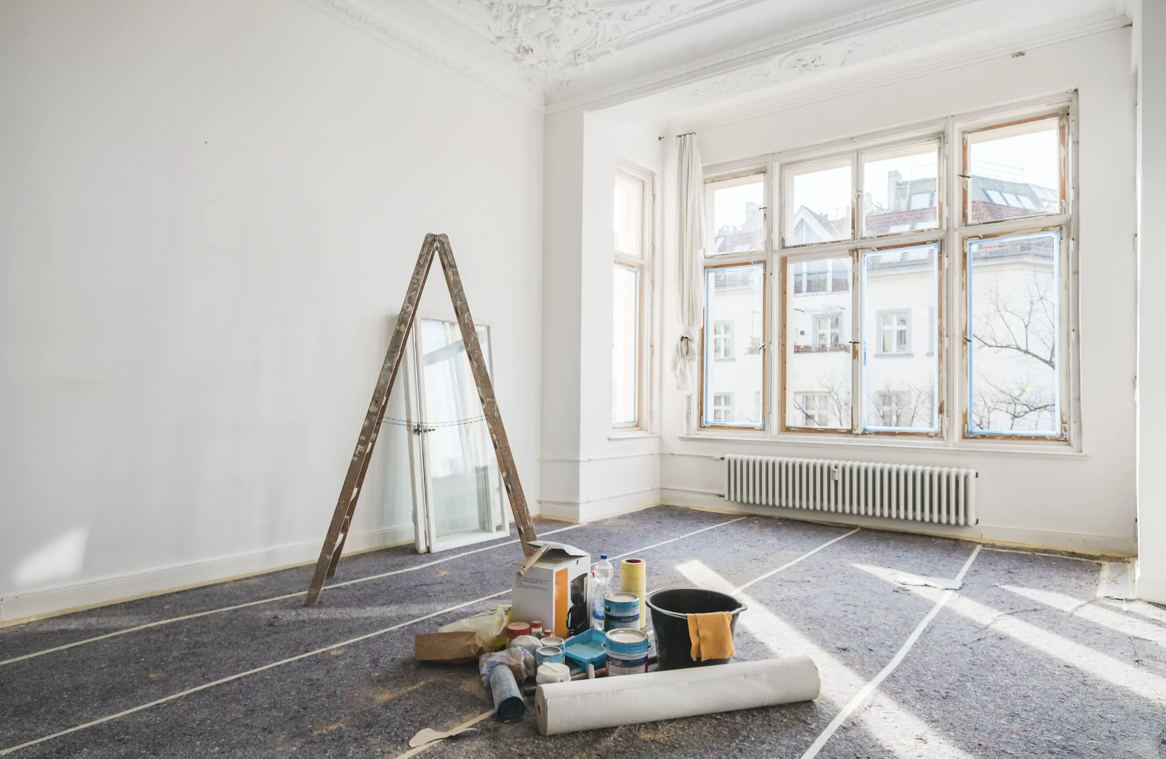 Huone remontin alla, jossa on maalaustarvikkeita, tikkaat ja suuret ikkunat. Lattialla on suojapeite. Huone on valoisa ja seinät ovat valkoiset.