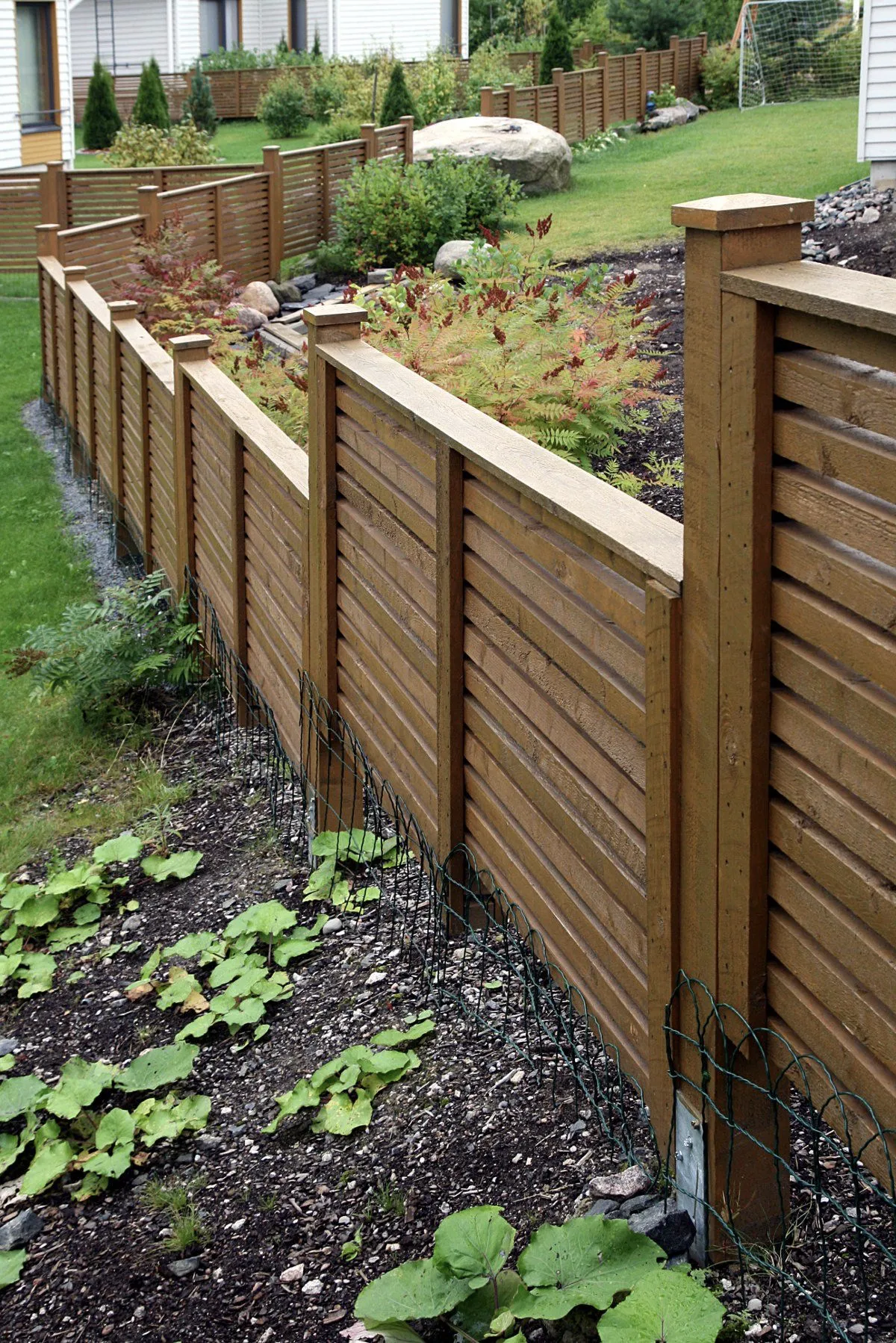 Ruskea puuaita erottaa puutarhan, jossa on vihreitä kasveja ja nurmikkoa, aurinkoisessa säässä.