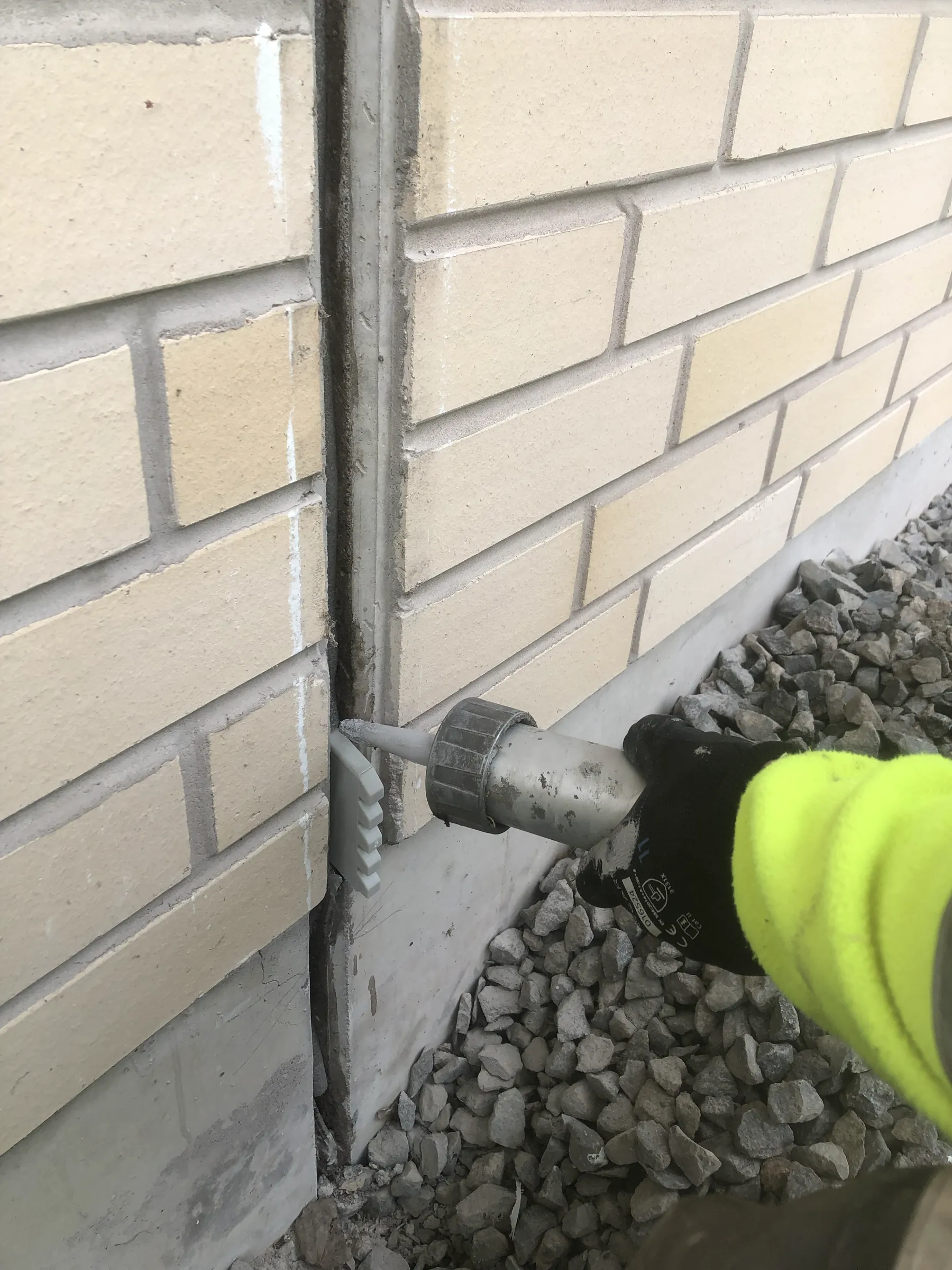 Kuva näyttää henkilön tiivistämässä rakennuksen seinän rakoa tiivistyspistoolilla, jossa on keltainen hiha ja musta käsine. Seinä on vaalean beige tiiliseinä, ja maassa on harmaita kiviä.
