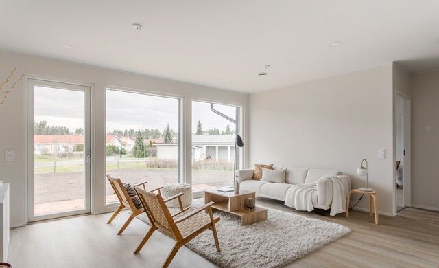 Valoisa olohuone, jossa on suuret ikkunat, vaalea sohva, puinen sohvapöytä ja kaksi rottinkituolia pehmeällä matolla.