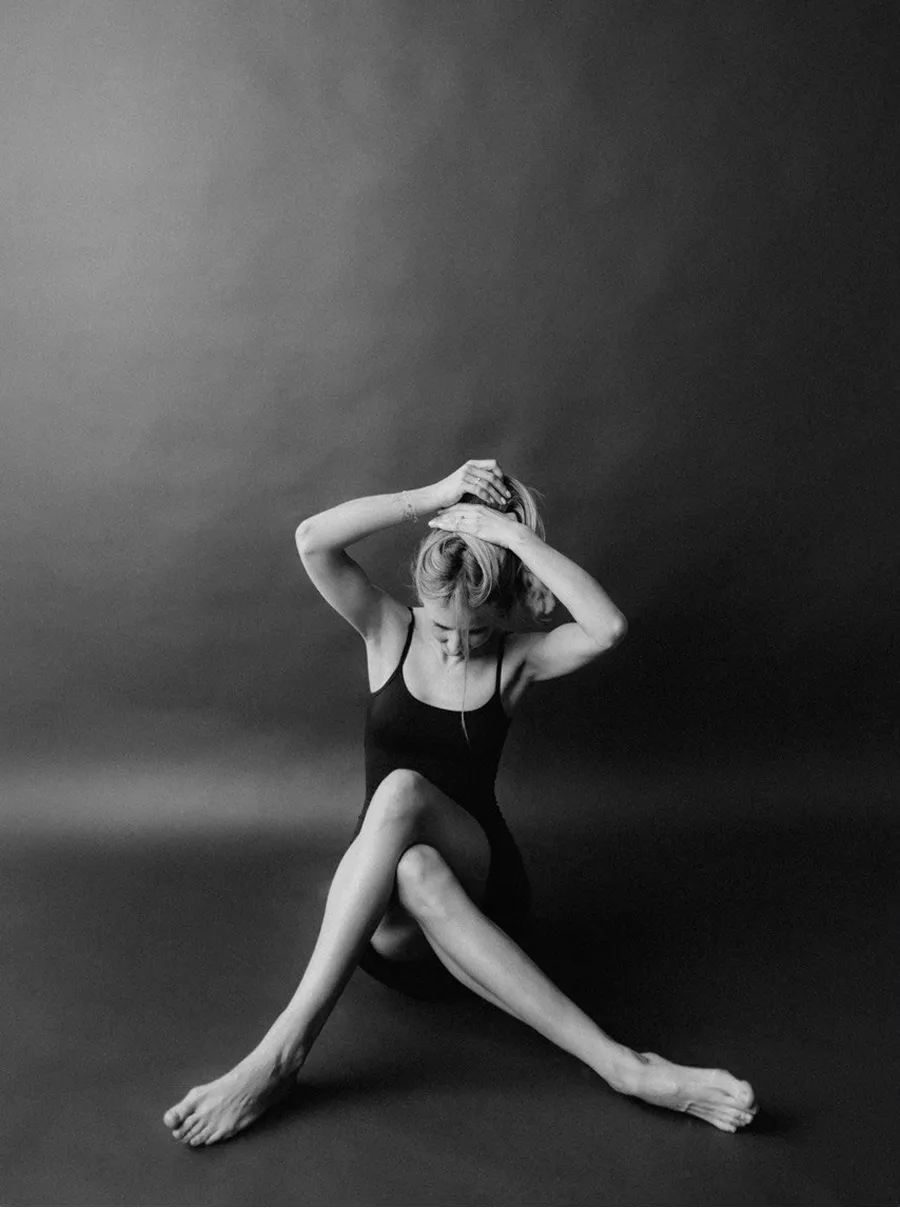 Жінка в чорному боді сидить на підлозі зі схрещеними ногами, піднімаючи руки до волосся на темному фоні.