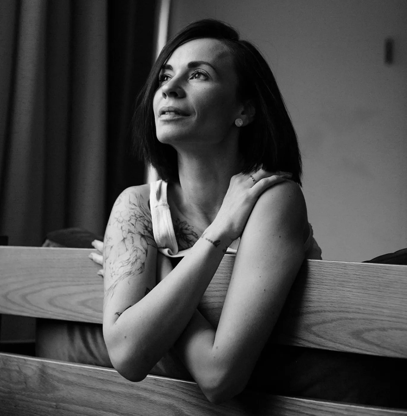 Жінка з коротким темним волоссям і тату на плечі, яка задумливо дивиться вгору, спираючись на дерев’яну спинку ліжка.