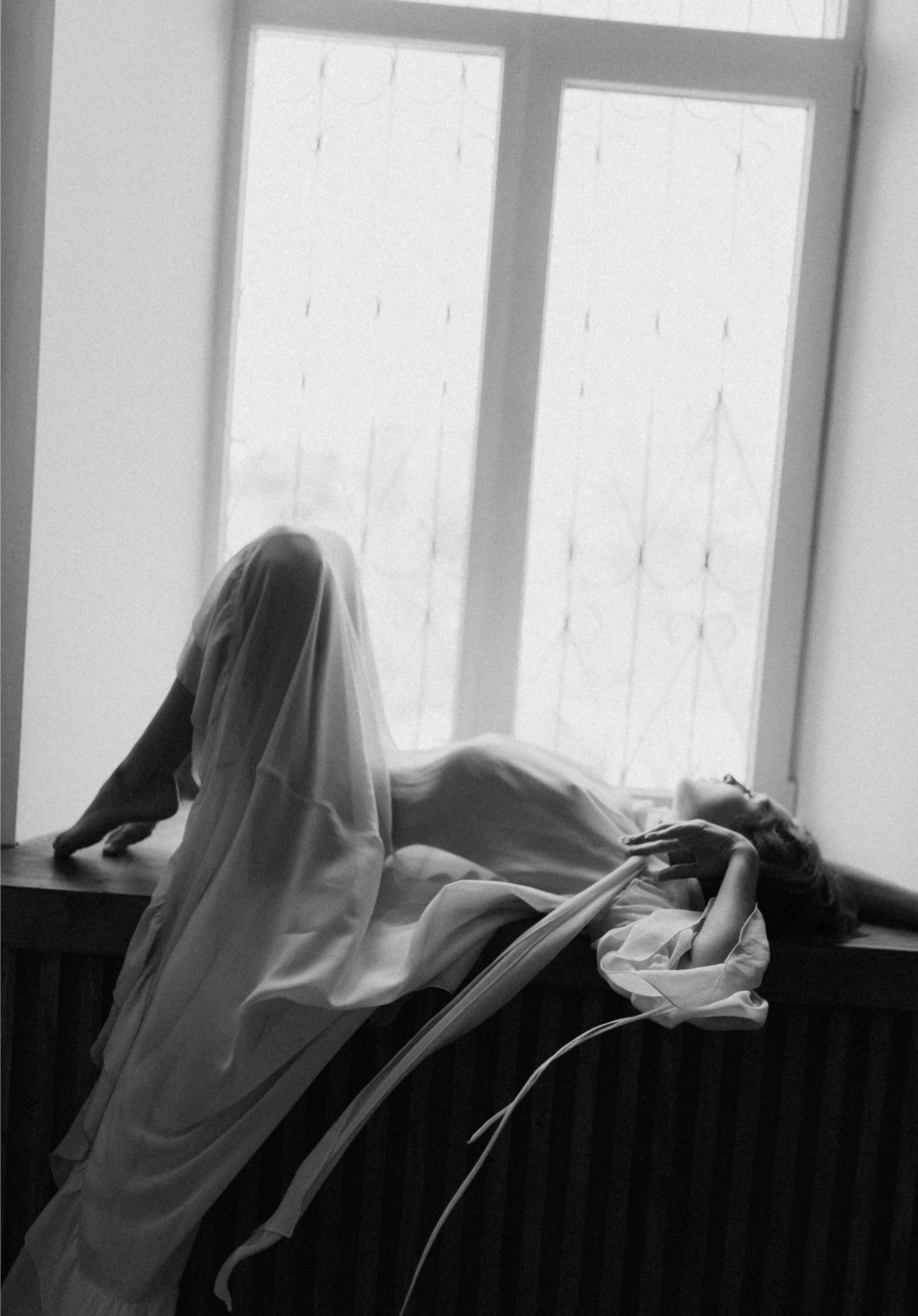 Жінка в довгій сукні лежить на віконній підвіконні, витягнувши руки і ноги, в монохромному тоні.