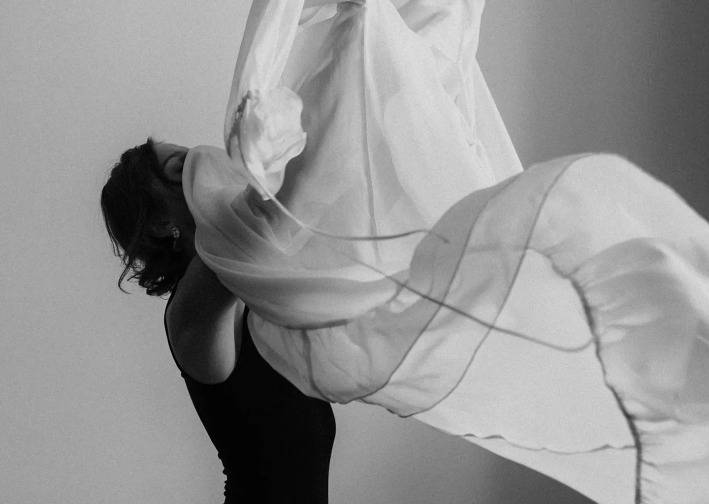 Жінка в чорній сукні кидає в повітря легку прозору тканину в чорнобілому фото.