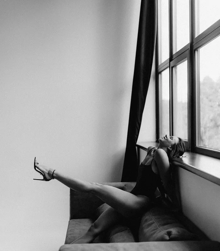 Жінка у чорному боді і високих підборах позує, витягнувши ногу, сидячи на дивані біля великого вікна.